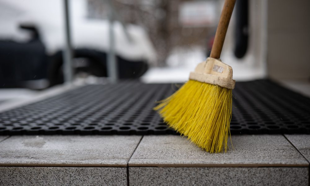 Shot of a wooden-handled broom, standing on the ground in front of a black door mat