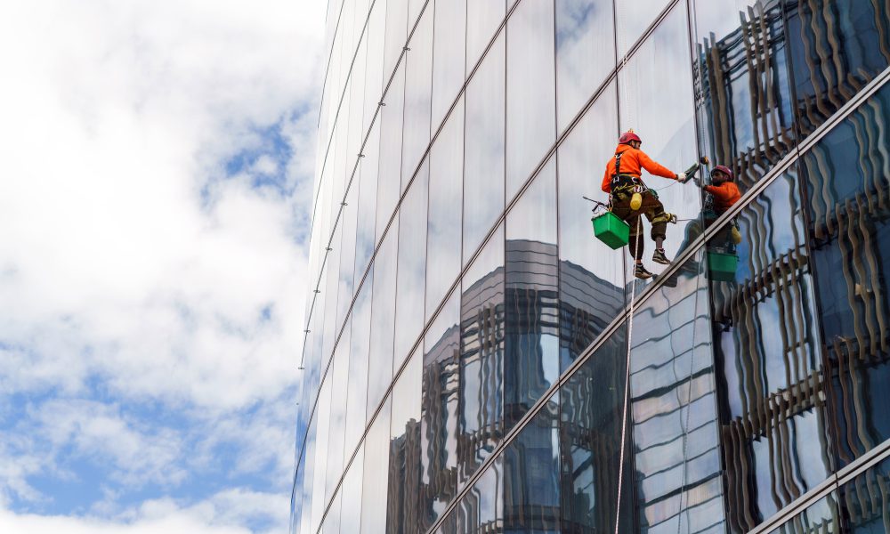 Worker in safety gear clean the windows of a tall glass building, suspended high above the ground.